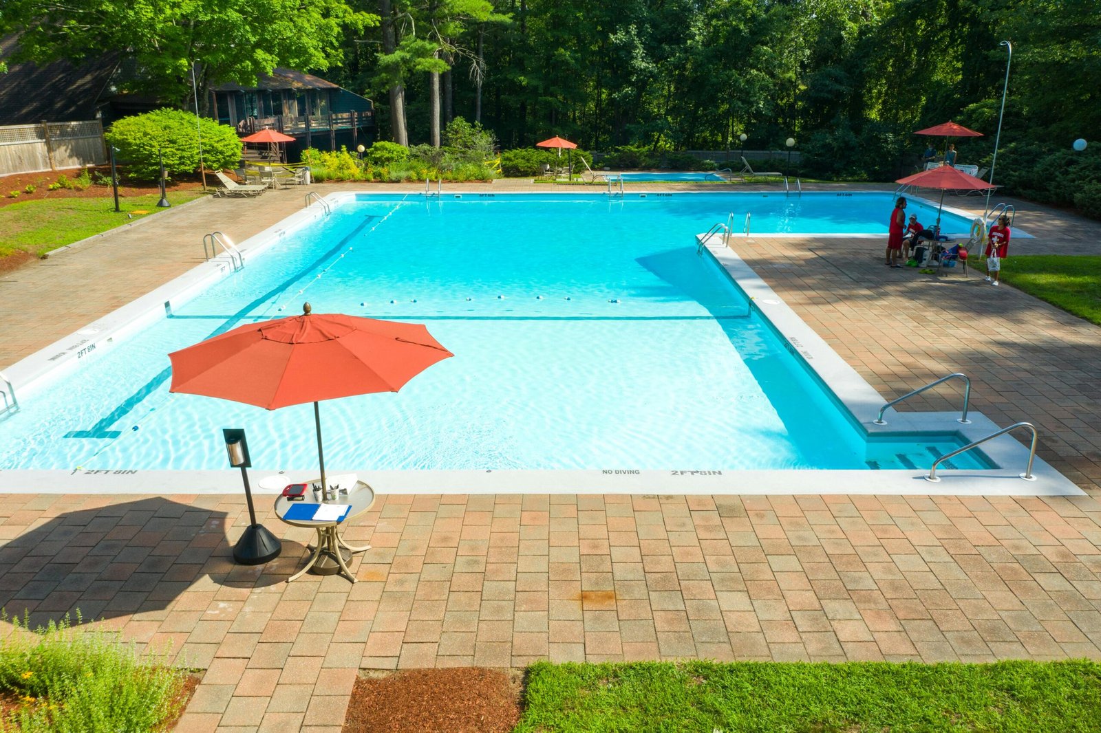A spacious outdoor swimming pool with red umbrellas and lounge area, taken on a sunny summer day.