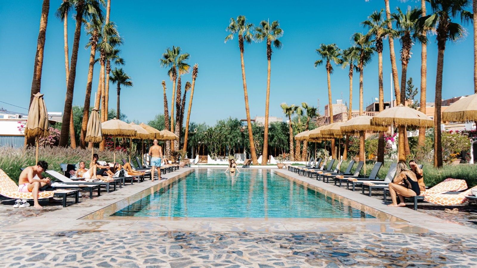 People relaxing by a palm-lined pool at a sunny resort with parasols and loungers.